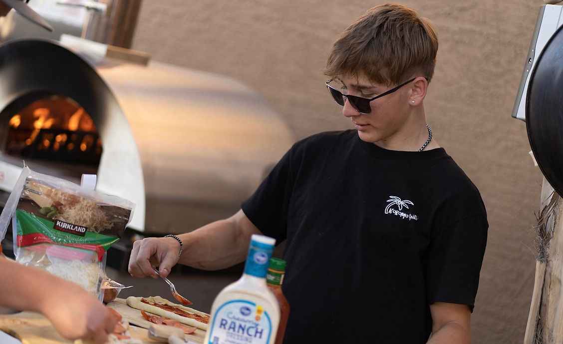 Outdoor pizza cooking at a wood-fired oven, preparing toppings near the opening