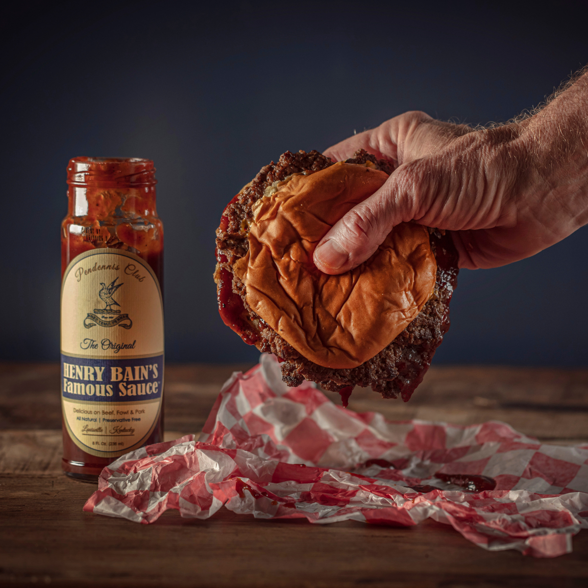 Sauce bottle with label beside a hand holding a sandwich