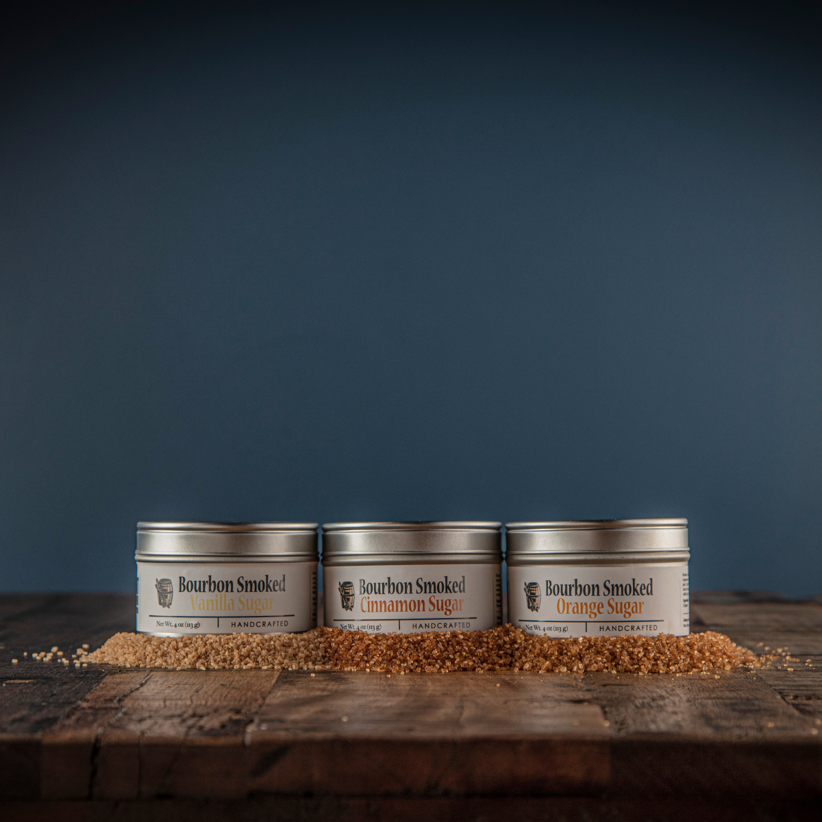 Three small metal tins of bourbon smoked sugar with orange, cinnamon, and vanilla labels