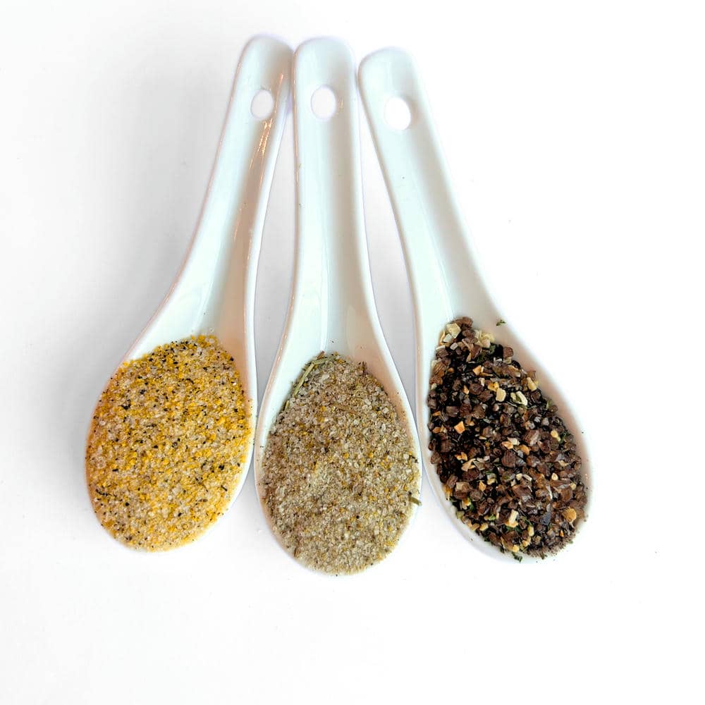 Three white measuring spoons filled with infused sea salt blends