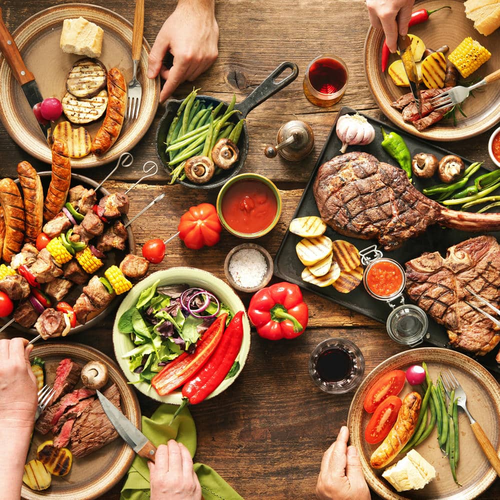 Mixed grilled meal spread on a wooden table with vegetables, bread, and steak