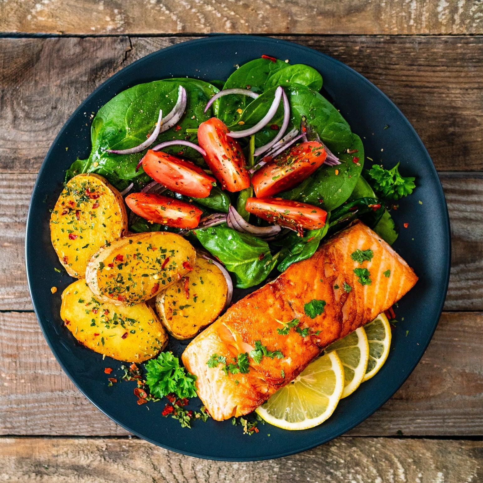 Seasoned salmon with lemon, salad greens, sliced onions and potatoes on a plate.