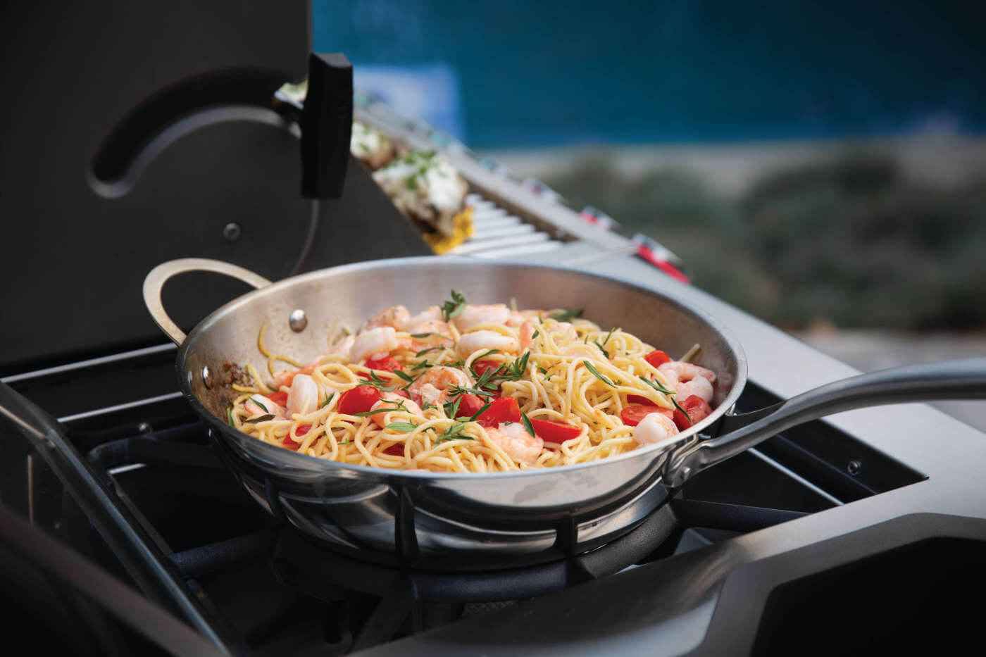 Side burner skillet with spaghetti and vegetables on a gas grill grate