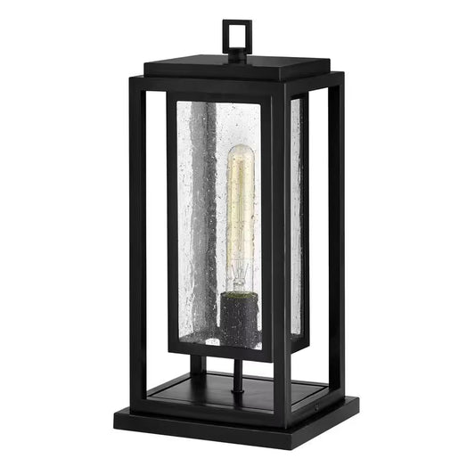 Black outdoor wall lantern with single clear glass panel and glowing bulb inside