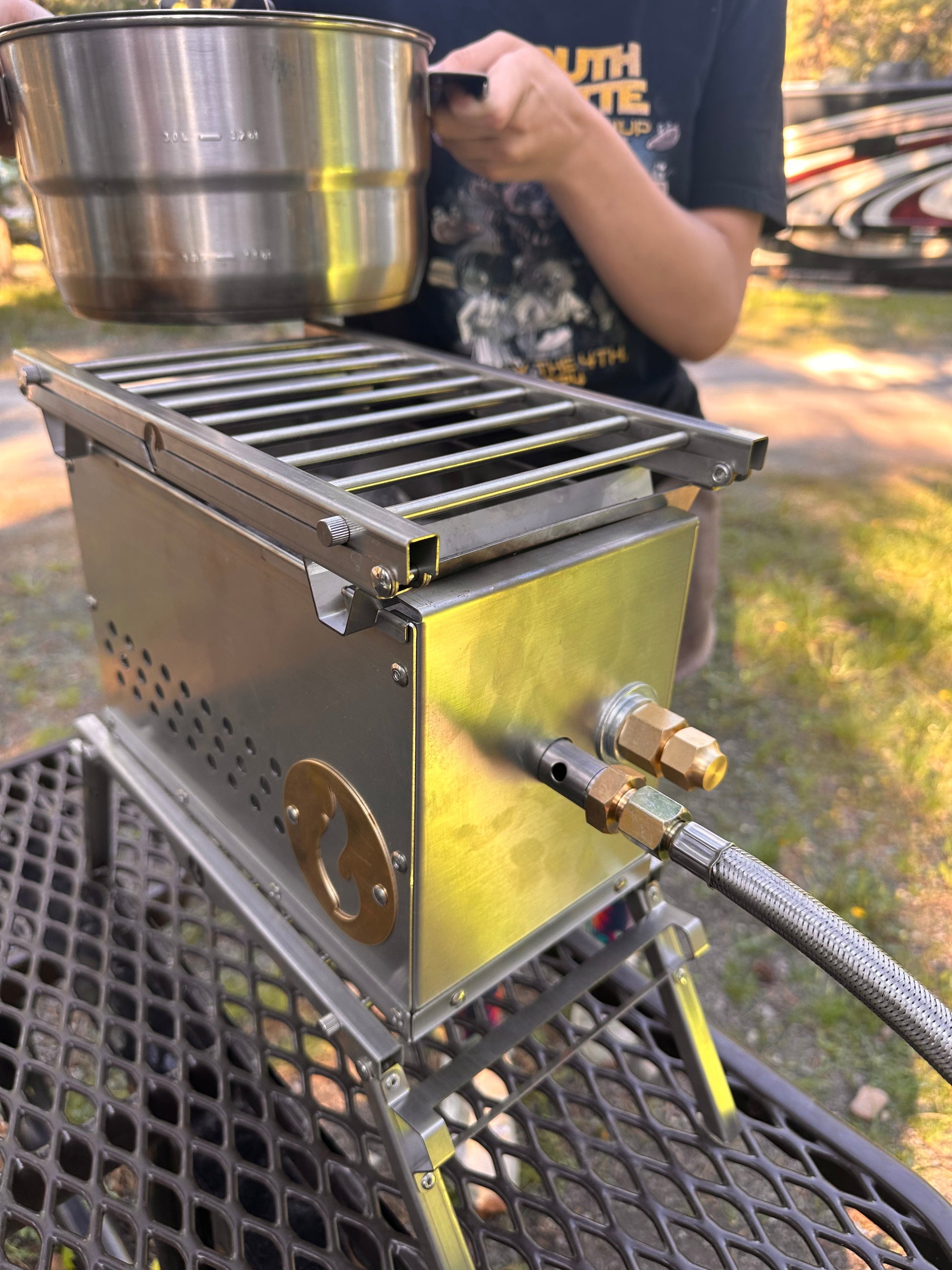 Stainless steel campfire grill with grates, side burner controls, and a pot on top